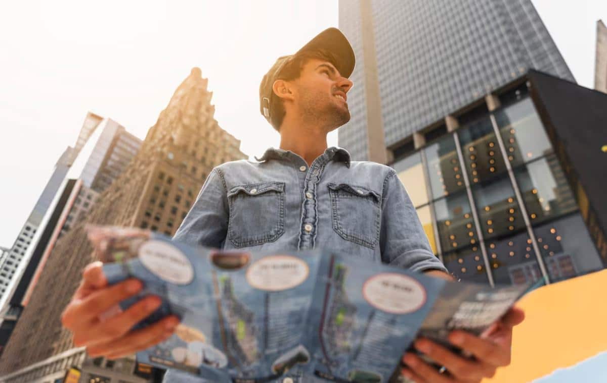 Man in a denim jacket looking up while holding maps, standing in an urban setting with skyscrapers.
