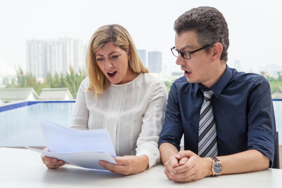 Two business professionals, a man and a woman, look shocked while reviewing documents at a table with cityscape in the background.