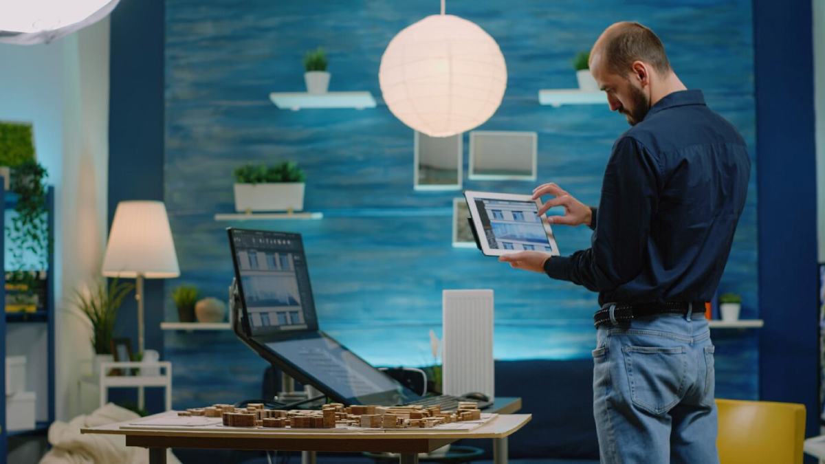 Man examining an architectural design on a tablet in a stylish, modern office with a laptop and model buildings on a table.