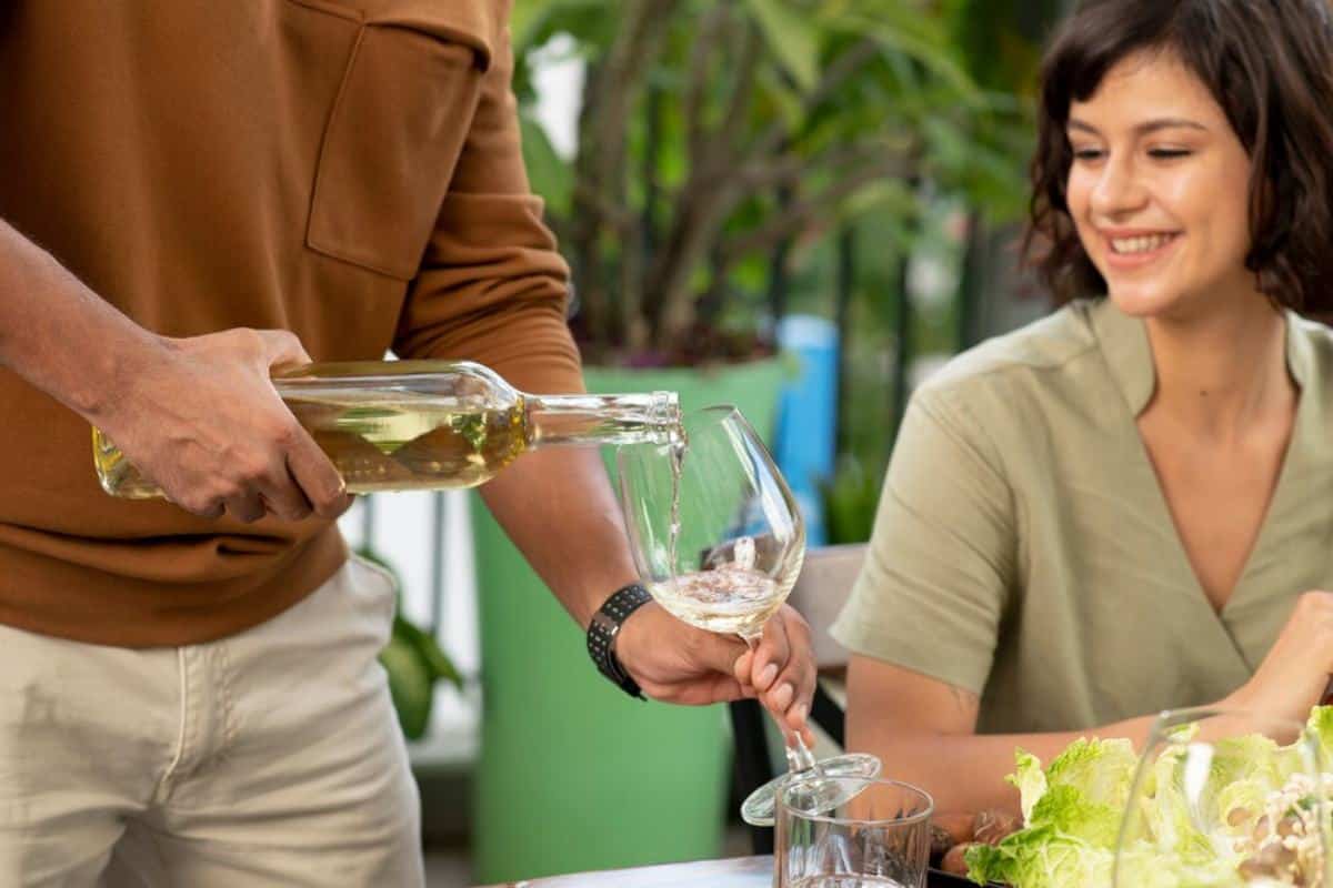 A hand pours white wine into a glass while a dining table adorned with fresh greens